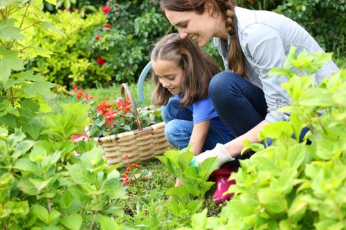 A woman and a young girl are engaged in outdoor gardening in a lush, well-maintained garden. The woman, dressed in a light grey top and blue jeans, is kneeling behind the girl, who is wearing a blue t-shirt and dark pants. They are both smiling and focused on planting or tending to plants. The garden features a vibrant mix of greenery, including leafy shrubs, ground cover plants, and flowering bushes with red blossoms. In the foreground, broad green leaves partially obscure the scene, while a wicker basket is placed on the ground, containing garden tools or plants. The background includes a variety of shrubs and small trees, creating a dense, natural environment typical of a suburban garden in Catford, London, or the surrounding area. The lighting suggests a clear day, with natural sunlight highlighting the fresh, lively colours of the garden. This scene reflects gardening activities such as planting, weeding, or nurturing outdoor plants, consistent with services offered by Lawn Mowing Catford and other garden maintenance providers in the local area.