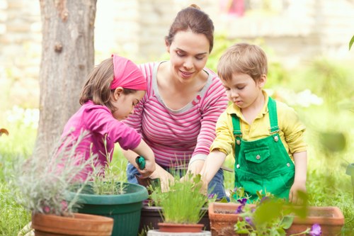A woman with dark hair tied back, wearing a pink and white striped long-sleeved top, is engaged in gardening activities outdoors in a residential garden. She is surrounded by two young children, one girl wearing a pink headband and a pink jumper, and a boy in a yellow shirt with green overalls, who are assisting her with planting in various pots and flower beds. The garden features a neatly maintained lawn area, with a large tree trunk visible on the left, and a low brick wall or fence in the background. Soil, small plants, and flowering blooms are arranged across the garden space, some in terracotta pots and others directly in the ground. The scene is illuminated by natural daylight, suggesting a mild, clear weather day, creating a warm and inviting outdoor environment suitable for gardening and family outdoor activities. The image conveys pruning, planting, and outdoor care, reflecting professional gardening and landscaping services offered by Lawn Mowing Catford and other local providers.