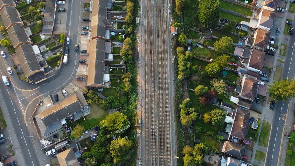 An aerial view of a residential street with terraced houses and neatly maintained back gardens. The gardens feature well-trimmed lawns, flower beds, and various trees and shrubs. Some gardens have pat